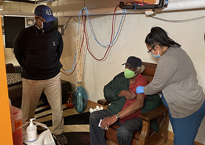 picture of Delroy King receiving COVID-19 vaccine from Emmaly Stallings as Mariners broadcaster Dave Sims looks on